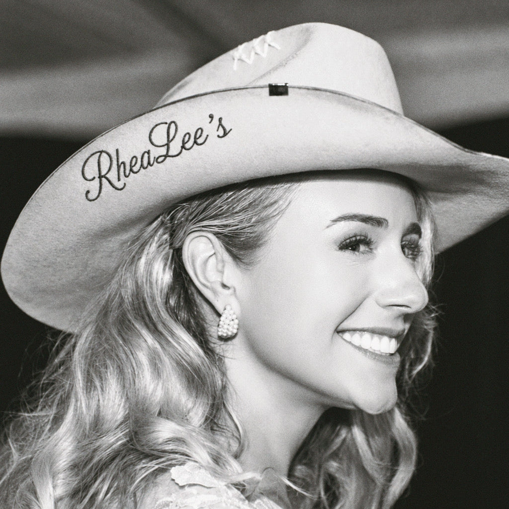A smiling woman in profile wearing a light-colored cowboy hat with the text 'RheaLee's' embroidered on the brim, shown in black and white.